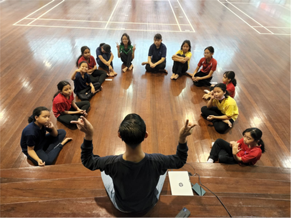 An adult instructor gestures with both hands toward a semicircle of children seated on a gym floor.